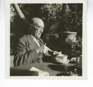 portrait photographique d’André Gide, avec lunettes, attablé pour un petit-déjeuner en plein air, beurrant une tartine, octobre 1936