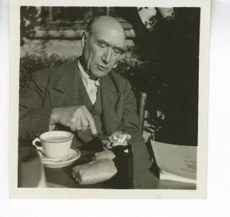 portrait photographique d’André Gide, attablé pour un petit-déjeuner en plein air, beurrant une tartine tout en parlant, octobre 1936