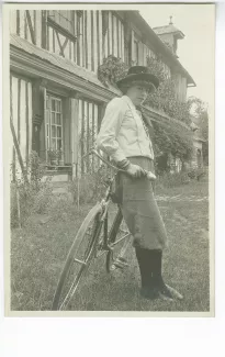 portrait photographique de Lucie Delarue-Mardrus, tenant son vélo, devant une maison à colombage