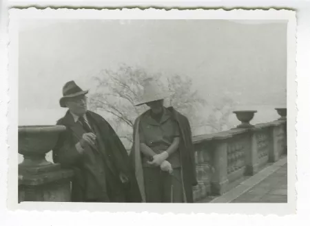 photographie d’André Gide, adossé à un balcon de pierre dans un lieu public, avec chapeau et lunettes, et une femme inconnue, portant un grand chapeau, octobre 1932