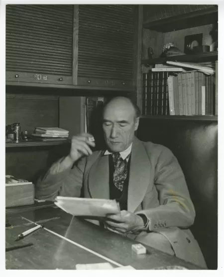 portrait photographique d’André Gide, avec lunettes, assis à son bureau, lisant, cigarette dans la main droite, au Vaneau