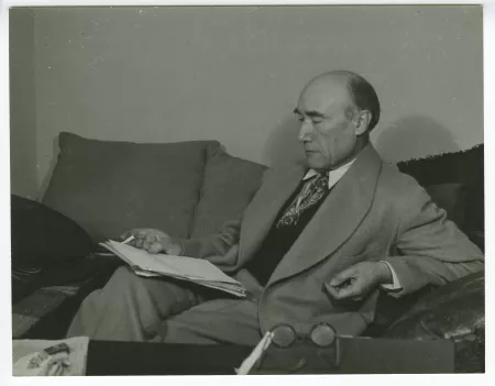 portrait photographique d’André Gide, assis sur un sofa, lisant, une cigarette dans la main droite, au Vaneau