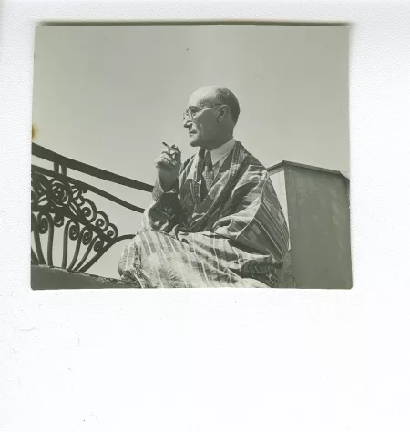portrait photographique d’André Gide, avec lunettes, de profil, assis sur le balcon du Vaneau, habillé en kimono-djellaba, une cigarette dans la main droite, juin 1935