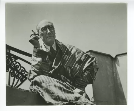 portrait photographique d’André Gide, avec lunettes, de face, assis sur le balcon du Vaneau, habillé en kimono-djellaba, une cigarette dans la main droite, juin 1935