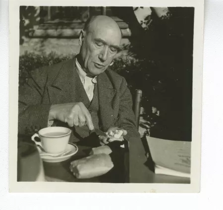 portrait photographique d’André Gide, attablé pour un petit-déjeuner en plein air, beurrant une tartine tout en parlant, octobre 1936