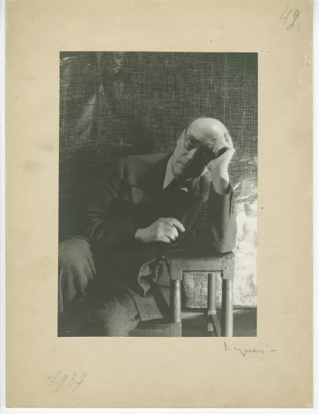 portrait photographique d’André Gide, avec lunettes, assis, accoudé à un petit escabeau, la tête appuyée sur la main gauche