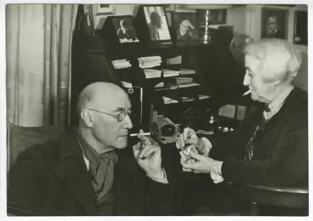 photographie d’André Gide, avec lunettes et foulard, se faisant allumer une cigarette par Maria Van Rysselberghe, assise à son bureau-secrétaire, rue Vaneau