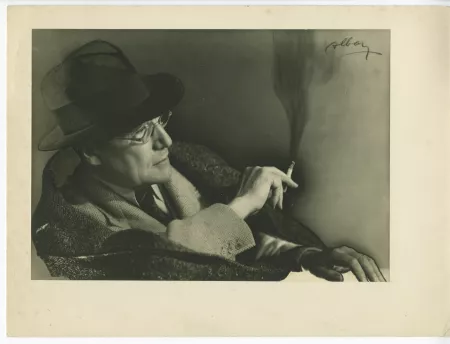 portrait photographique en buste d’André Gide, avec chapeau et lunettes, assis, de profil à droite, une cigarette dans la main droite, la gauche posée sur une table, mars 1939