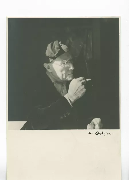portrait photographique d’André Gide, avec lunettes et bonnet, de ¾ à droite, assis à son bureau, fumant, au Vaneau