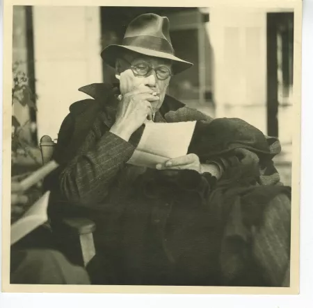 photographie d’André Gide, avec lunettes et chapeau lisant une page volante de livre, et fumant, en extérieur