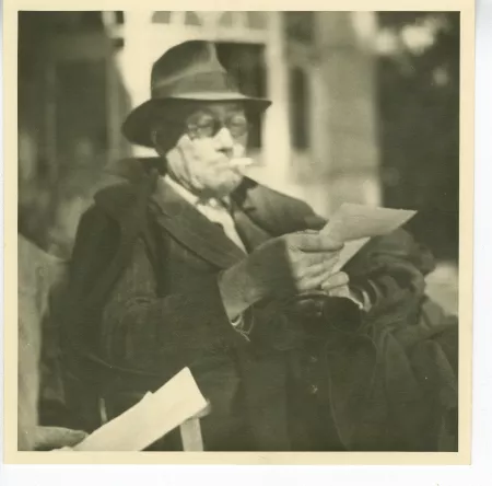 photographie d’André Gide, avec lunettes et chapeau lisant une page volante de livre, et fumant, en extérieur, château de Bussigny, août 1946