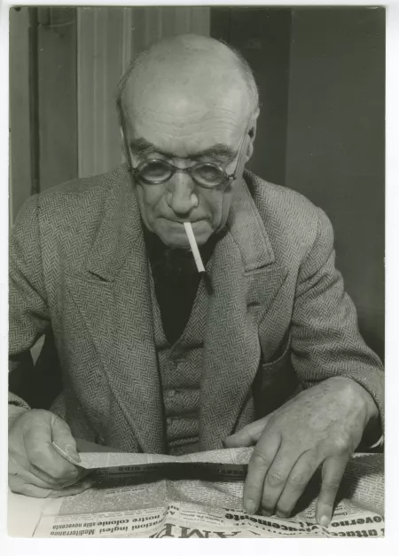 portrait photographique d’André Gide, avec lunettes, lisant le journal italien La Stampa, une cigarette à la bouche, novembre 1947