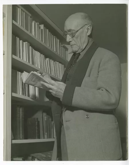 portrait photographique d’André Gide, avec lunettes, lisant un livre de Georges Simenon, debout à côté d’une bibliothèque, chez Richard Heyd, hiver 1947