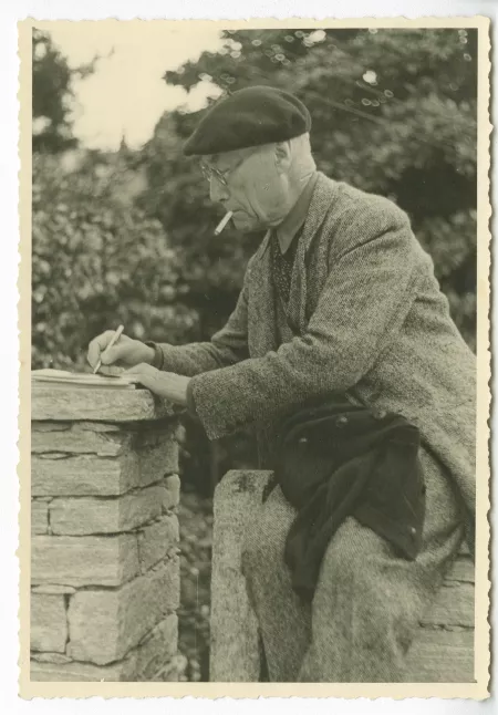 portrait photographique d’André Gide, avec lunettes et béret, de profil, écrivant sur un muret, en extérieur, avril 1947