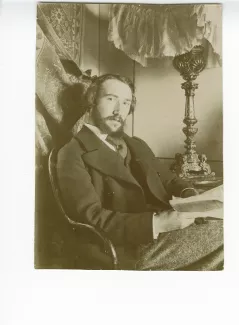 portrait photographique d’André Gide assis, à la barbe en double pointe, un livre entre les mains, dans sa chambre du Royal-Hôtel, février-mars 1895