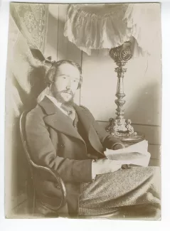 portrait photographique d’André Gide assis, à la barbe en double pointe, un livre entre les mains, dans sa chambre du Royal-Hôtel, février-mars 1895