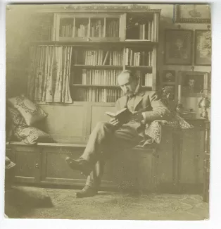 photographie d’André Gide, avec moustache, assis, lisant sur un banc de bois dans une pièce de la maison de la rue Laugier, chez Maria et Théo Van Rysselberghe