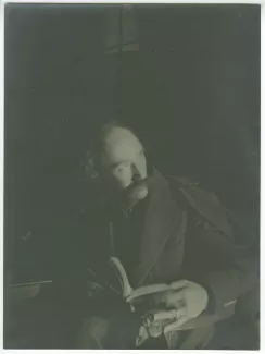 portrait photographique d’André Gide, avec moustache, assis, tenant un livre, une bague à la main gauche, dans un intérieur sombre