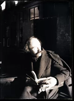 portrait photographique d’André Gide, avec moustache, assis, tenant un livre, une bague à la main gauche, dans un intérieur sombre