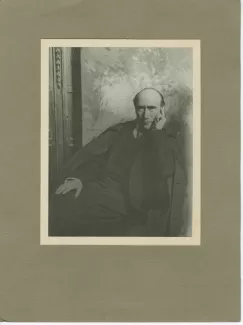 portrait photographique d’André Gide, assis, de face, devant une toile peinte, collé sur carton gris