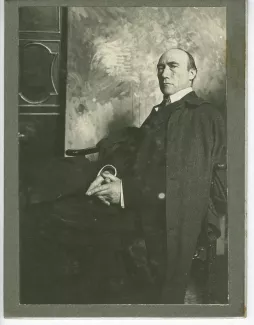 portrait photographique d’André Gide, assis, de ¾, mains jointes, devant une toile peinte