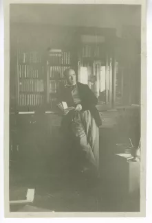 photographie d’André Gide, debout, livre à la main, devant une bibliothèque, chez les Mayrisch à Dudelange, Pâques 1919