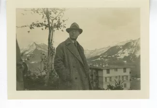 photographie d’André Gide, avec lunettes chapeau, mains dans les poches, à l'extérieur devant un paysage de montagnes, décembre 1933