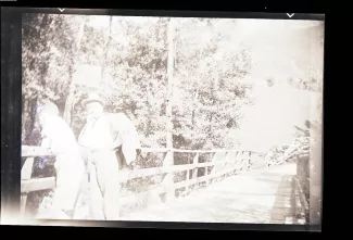 photographie d'André Gide, avec lunettes et chapeau, accoudé à côté d’un adolescent, sur un pont