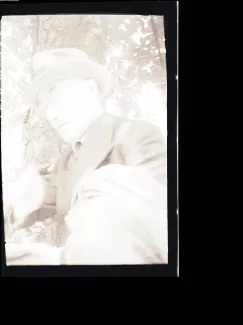 portrait photographique en buste d'André Gide, avec lunettes, chapeau et veste au bras, une cigarette dans la main droite et lisant, août 1934