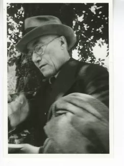 portrait photographique en buste d'André Gide, avec lunettes, chapeau et veste au bras, une cigarette dans la main droite et lisant, août 1934