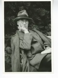 portrait photographique d'André Gide, avec lunettes et chapeau, adossé à une chaise et tourné vers l’objectif, la main droite sur le menton, la gauche tenant un livre, août 1934