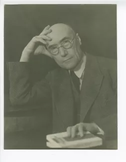 portrait photographique en buste d’André Gide, avec lunettes, regardant l’objectif, la main droite sur le front et la gauche tenant un livre, août 1934