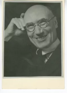 portrait photographique en buste d’André Gide, avec lunettes, la main droite sur le front, riant, août 1934