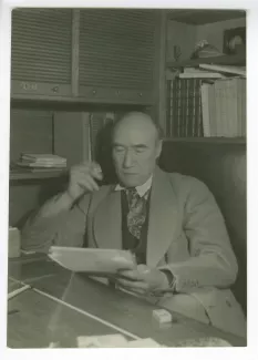 portrait photographique d’André Gide, avec lunettes, assis à son bureau, lisant, cigarette dans la main droite, au Vaneau