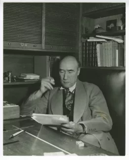 portrait photographique d’André Gide, avec lunettes, assis à son bureau, lisant, cigarette dans la main droite, au Vaneau