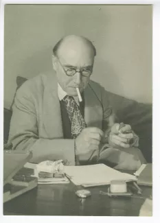 portrait photographique en buste d’André Gide, avec lunettes, assis à une table, lisant, en train d’allumer une cigarette, au Vaneau