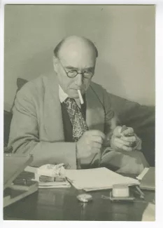 portrait photographique en buste d’André Gide, avec lunettes, assis à une table, lisant, en train d’allumer une cigarette, au Vaneau