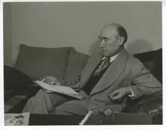 portrait photographique d’André Gide, assis sur un sofa, lisant, une cigarette dans la main droite, au Vaneau