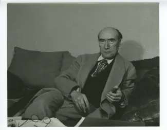 portrait photographique d’André Gide, assis sur un sofa, regardant l’objectif, une cigarette dans la main droite et un briquet dans la gauche, au Vaneau