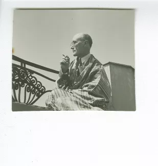 portrait photographique d’André Gide, avec lunettes, de profil, assis sur le balcon du Vaneau, habillé en kimono-djellaba, une cigarette dans la main droite, juin 1935