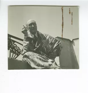 portrait photographique d’André Gide, avec lunettes, de face, assis sur le balcon du Vaneau, habillé en kimono-djellaba, une cigarette dans la main droite, juin 1935