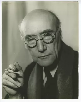 portrait photographique en buste d’André Gide, avec lunettes, de face, regardant vers le bas, une cigarette dans la main droite