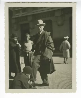 photographie en pied d’André Gide, debout, avec lunettes et chapeau, se faisant cirer les chaussures dans une rue d'Alger, 15 février 1936