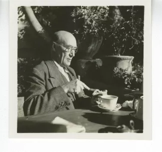 portrait photographique d’André Gide, avec lunettes, attablé pour un petit-déjeuner en plein air, beurrant une tartine, octobre 1936
