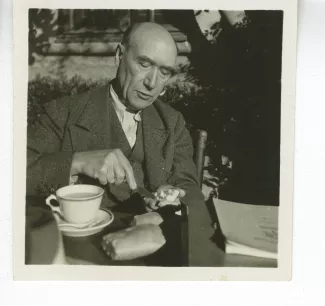 portrait photographique d’André Gide, attablé pour un petit-déjeuner en plein air, beurrant une tartine tout en parlant, octobre 1936