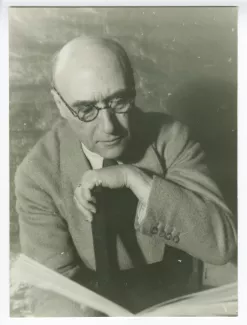 portrait photographique en buste d’André Gide, avec lunettes, de face, lisant, le bras gauche sur le dossier d'une chaise