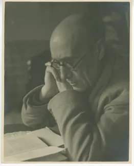 portrait photographique en buste d’André Gide, avec lunettes, lisant à un bureau, les mains jointes