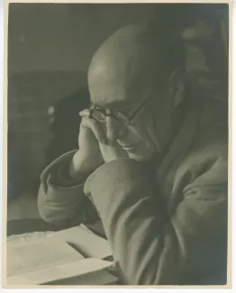 portrait photographique en buste d’André Gide, avec lunettes, lisant à un bureau, les mains jointes