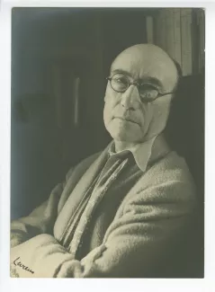 portrait photographique en buste d’André Gide, avec lunettes et foulard fleuri, regardant l’objectif, les bras croisés, adossé à une bibliothèque, au Vaneau