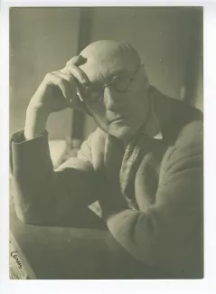 portrait photographique en buste d’André Gide, avec lunettes et foulard fleuri, attablé, regardant l’objectif, la tête appuyée sur la main droite, au Vaneau